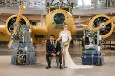 A bride and groom posing for a wedding photo in front of a large, vintage yellow airplane inside an aviation museum, surrounded by elegant floral arrangements and vintage-inspired decor.